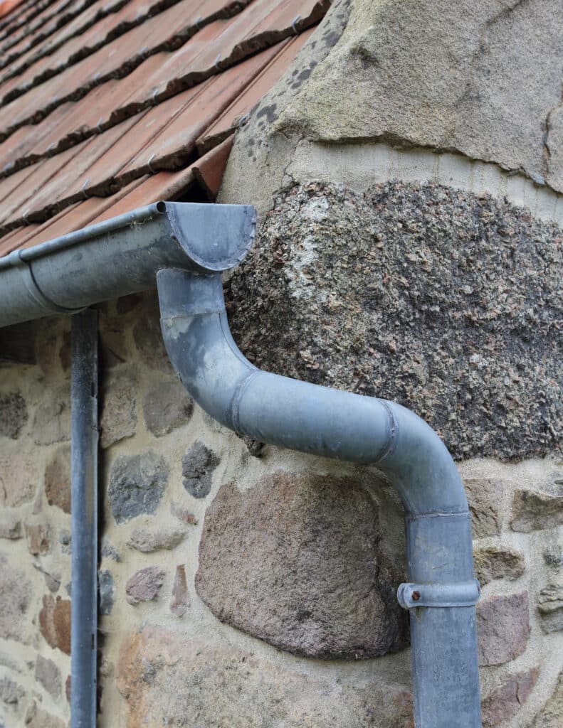 Gouttière en Zinc et Descente Pluviale sur Mur en Pierre Gouttière en zinc grise avec descente d'eau pluviale courbée sur un mur en pierre naturelle et toit en tuiles.