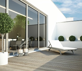 Terrasse moderne en bois avec chaise longue, baies vitrées et arbustes décoratifs sous un ciel bleu.
